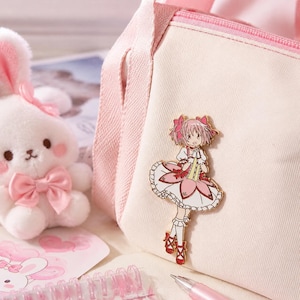 Puede incluir: Un bolso de lona rosa claro con un pin de esmalte dorado de un personaje de anime con cabello rosa, que viste un vestido blanco y rosa con detalles rojos. El personaje lleva lazos rosas en el cabello y zapatos rojos con cordones.