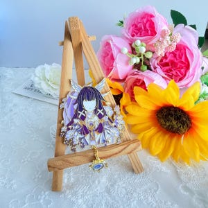 Puede incluir: Un broche de esmalte en tono dorado que presenta un personaje estilizado con cabello morado oscuro, ropa blanca y morada, y alas, acentuado por un pequeño dije colgante. El broche se exhibe en un pequeño caballete de madera.