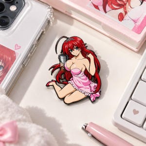 May include: A metal enamel pin featuring an anime character with long red hair, wearing a small silver tiara and a light pink slip dress. The character is depicted in a kneeling pose, holding a small silver trophy cup in one hand.