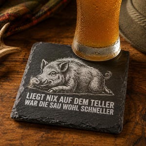 Könnte beinhalten: Ein quadratischer Untersetzer aus schwarzem Schiefer mit einer eingravierten Wildschwein-Illustration und dem weißen Schriftzug "LIEGT NIX AUF DEM TELLER WAR DIE SAU WOHL SCHNELLER", auf dem ein Glas mit goldenem Bier steht.