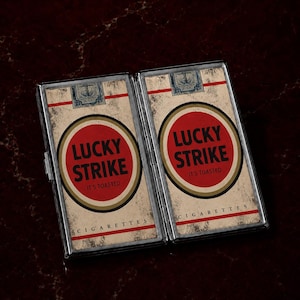 Może przedstawiać: Prostokątna metalowa papierośnica z nadrukiem w stylu vintage Lucky Strike po obu stronach. Grafika zawiera czerwone koło z napisem Lucky Strike i It's Toasted, niebieski znaczek oraz słowo Cigarettes czarnymi literami na beżowym tle.
