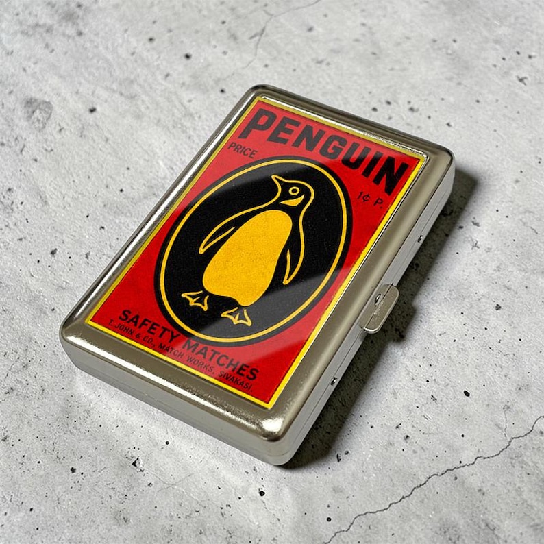 Puede incluir: Una caja rectangular de metal plateado con una etiqueta de estilo vintage sobre fondo rojo, que presenta un &oacute;valo negro con una ilustraci&oacute;n de un ping&uuml;ino amarillo. El texto dice PENGUIN, PRICE, 1c P., SAFETY MATCHES, T. JOHN & CO., MATCH WORKS, SIVAKASI.