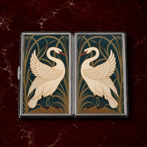 May include: A silver cigarette case with a decorative design featuring two white swans facing each other against a teal background with gold and brown accents. The swans have red beaks and are surrounded by stylized reeds.