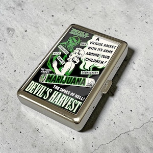 Puede incluir: Una pitillera rectangular de metal con un gráfico de estilo vintage en tonos verdes y negros. El diseño incluye una figura de diablo, una mujer y el texto: DEVIL'S HARVEST, A VICIOUS RACKET WITH IT'S ARMS AROUND YOUR CHILDREN, MARIJUANA.