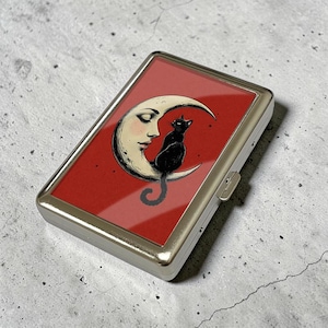 Puede incluir: Un estuche de cigarrillos de metal plateado con un fondo rojo que presenta una luna creciente con el rostro de una mujer y un gato negro. El gato está sentado en la luna. El estuche tiene forma rectangular.