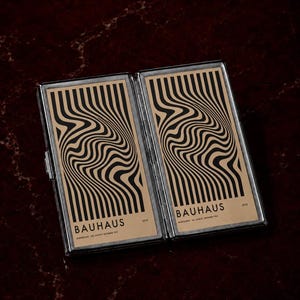 May include: A rectangular, hinged metal case featuring two identical panels with a tan background and black, wavy, vertical line patterns. Each panel displays the text "BAUHAUS" at the bottom, with smaller text "1919" and "AUSSTELLUNG JULI, AUGUST, SEPTEMBER 1923".
