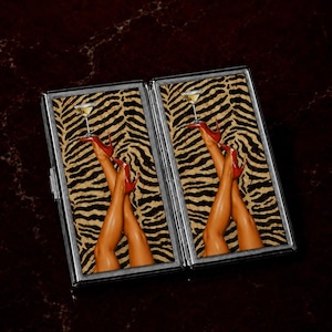 May include: A rectangular metal cigarette case featuring a hinged design. The interior panels display a repeating graphic of crossed legs wearing red high-heeled shoes, with one foot balancing a martini glass against a black and tan tiger print background.