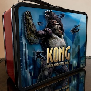 May include: A rectangular metal lunch box with a black handle and red sides. The front features a graphic of a giant ape with the text KONG THE 8TH WONDER OF THE WORLD, depicting the creature amidst a city skyline with airplanes flying around it.