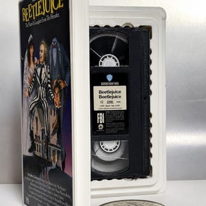 Può includere: Una custodia per VHS in miniatura in plastica bianca per il film Beetlejuice, con la locandina del film sulla copertina. All'interno, è visibile una cassetta VHS nera con un'etichetta che riporta la scritta Beetlejuice Beetlejuice. Una moneta d'argento è accanto.