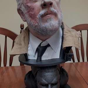 Puede incluir: Un busto detallado de una persona mayor con barba blanca, ojos azul brillante y cicatrices faciales, que viste una camisa blanca de cuello, corbata negra y chaqueta beige. La base presenta un rostro oscuro esculpido con acabado negro mate.