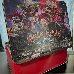 以下が含まれることがあります： 映画「Killer Klowns from Outer Space」のグラフィックプリントが施された赤い金属製のランチボックス。蓋には3人のピエロ、2人の人物、パトカー、アイスクリームトラックが描かれており、「Killer Klowns from Outer Space」という文字が記されています。