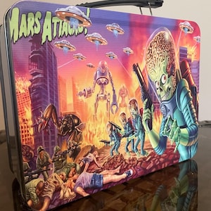 Puede incluir: Una lonchera rectangular de metal con un gráfico colorido de Mars Attacks!. La ilustración muestra una escena caótica con alienígenas de piel verde, platillos voladores y una ciudad en llamas. Un asa de plástico negro está fijada en la parte superior.