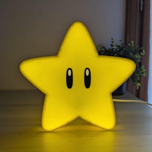 May include: A yellow star-shaped night light with black oval eyes. The light is glowing, and the star is sitting on a wooden surface. The light is plugged into an electrical outlet. The star is a popular video game character.