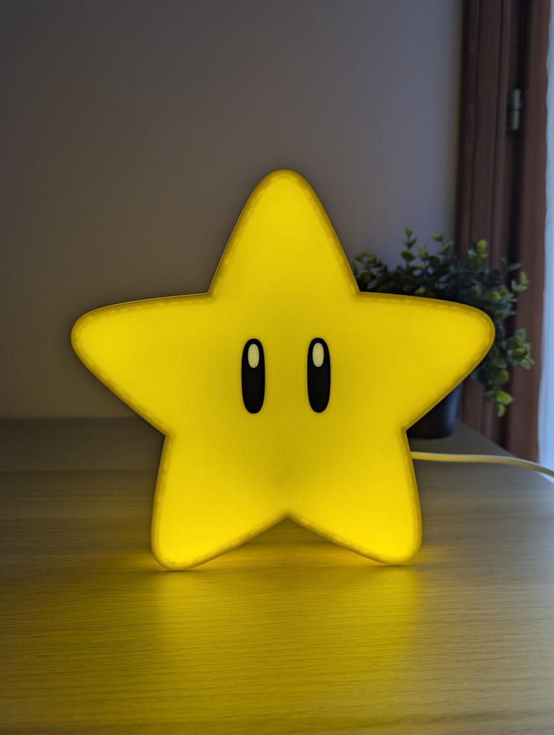 May include: A yellow star-shaped night light with black oval eyes. The light is glowing, and the star is sitting on a wooden surface. The light is plugged into an electrical outlet. The star is a popular video game character.