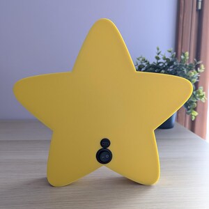 May include: A bright yellow star-shaped night light with a smooth surface. The light has a black on/off switch and a USB port on the back. It is sitting on a light-colored wooden surface.