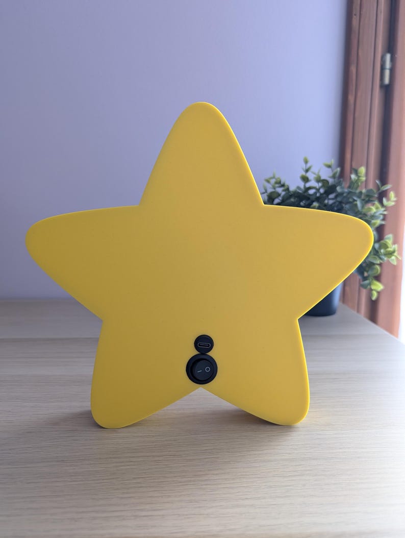 May include: A bright yellow star-shaped night light with a smooth surface. The light has a black on/off switch and a USB port on the back. It is sitting on a light-colored wooden surface.