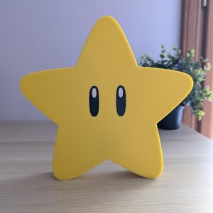 May include: A yellow star-shaped decorative item with black oval eyes. The star is a bright yellow color and sits on a light-colored wooden surface. The item is a stylized representation of a star, commonly seen in video games.