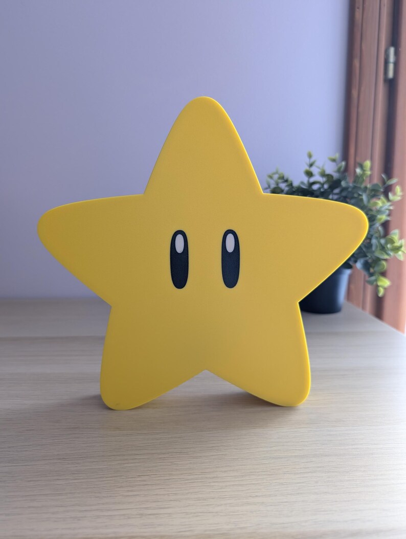 May include: A yellow star-shaped decorative item with black oval eyes. The star is a bright yellow color and sits on a light-colored wooden surface. The item is a stylized representation of a star, commonly seen in video games.