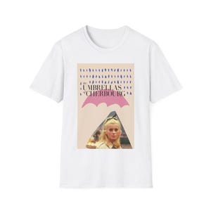 May include: White t-shirt with a graphic from the film "The Umbrellas of Cherbourg." The design features a pink umbrella, blue raindrops, and a woman's portrait. The text "The Umbrellas of Cherbourg" is printed above the umbrella.