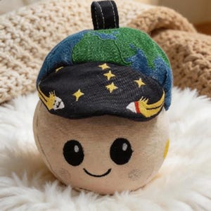 May include: A plush toy shaped like a smiling moon wearing a hat. The hat is blue and green, depicting the Earth, with a black brim featuring yellow stars and rockets. The toy has a black loop on top.