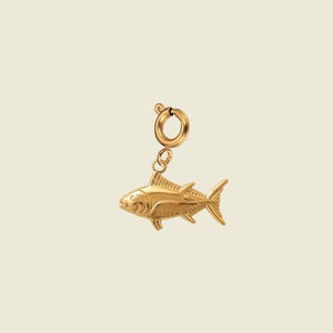 May include: A gold-toned metal charm in the shape of a tuna fish, featuring detailed engraved lines on the body, fins, and tail. The fish charm is attached to a circular spring ring clasp, allowing it to be easily connected to a chain or bracelet.