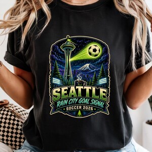 May include: Black t-shirt featuring a graphic design with the text "SEATTLE RAIN CITY GOAL SIGNAL SOCCER 2026". The design includes the Space Needle, a soccer ball, and a mountain range, with green and blue accents.