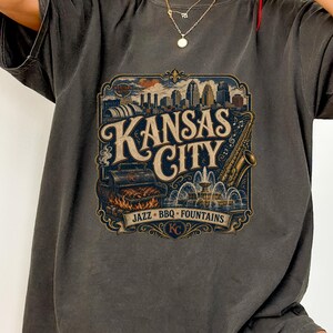 K&ouml;nnte beinhalten: Dunkelgraues T-Shirt mit einem Vintage-Grafikdesign von Kansas City. Das Design zeigt die Skyline der Stadt, einen Grill, ein Saxophon und einen Brunnen, mit den Worten "Kansas City" und "Jazz BBQ Fountains".