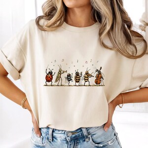 May include: A cream-coloured short-sleeve t-shirt featuring a graphic illustration of six insects playing musical instruments. The insects include a ladybug, a grasshopper, a beetle, a bee, and a mosquito, all depicted in a line with musical notes.