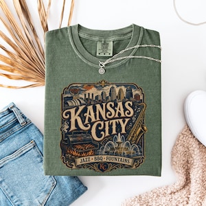 K&ouml;nnte beinhalten: Salbeigr&uuml;nes T-Shirt mit einem Vintage-Grafikdesign von "Kansas City". Das Design zeigt eine Skyline, ein Saxophon, einen Grill und einen Brunnen. Eine silberne Halskette liegt auf dem T-Shirt.