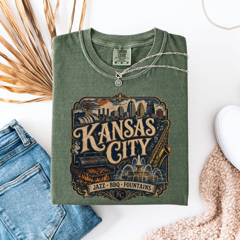 K&ouml;nnte beinhalten: Salbeigr&uuml;nes T-Shirt mit einem Vintage-Grafikdesign von "Kansas City". Das Design zeigt eine Skyline, ein Saxophon, einen Grill und einen Brunnen. Eine silberne Halskette liegt auf dem T-Shirt.