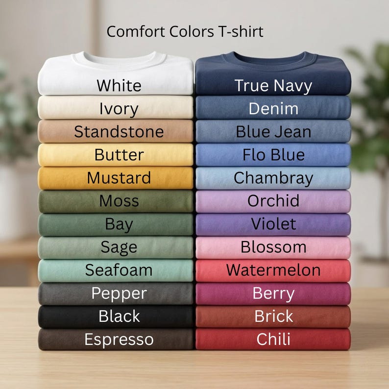 K&ouml;nnte beinhalten: Gestapelte Comfort Colors T-Shirts in verschiedenen Farben, darunter Wei&szlig;, Marineblau und Schwarz. Das Bild zeigt eine Reihe von Farbt&ouml;nen, von Pastellt&ouml;nen bis zu dunkleren Schattierungen, wobei jedes Shirt ordentlich gefaltet und mit seinem Farbnamen beschriftet ist.