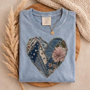 May include: A light blue t-shirt with a heart-shaped design made of denim patches and floral fabric. A gold necklace with a pendant rests on the shirt. The shirt is folded on a wooden surface.