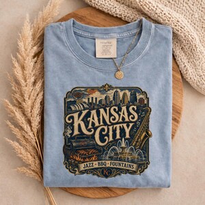 K&ouml;nnte beinhalten: Hellblaues T-Shirt mit einer Grafik der Skyline von Kansas City, einem Saxophon und den Worten "Kansas City Jazz BBQ Fountains". Eine goldene Halskette mit Anh&auml;nger liegt auf dem Shirt.