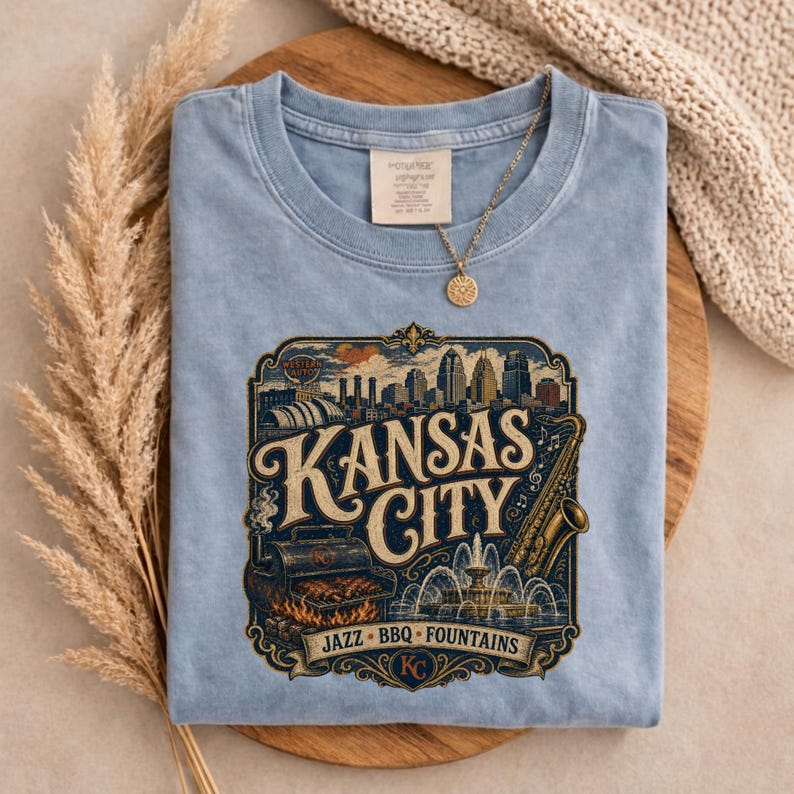 K&ouml;nnte beinhalten: Hellblaues T-Shirt mit einer Grafik der Skyline von Kansas City, einem Saxophon und den Worten "Kansas City Jazz BBQ Fountains". Eine goldene Halskette mit Anh&auml;nger liegt auf dem Shirt.