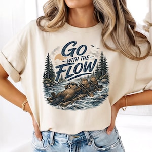 Peut inclure: Un t-shirt couleur crème avec une impression graphique représentant trois castors sur un radeau de bois dans une rivière. Le texte "Go WITH THE FLOW" est imprimé en bleu foncé au-dessus des castors. La scène inclut des pins, un soleil et des oiseaux.