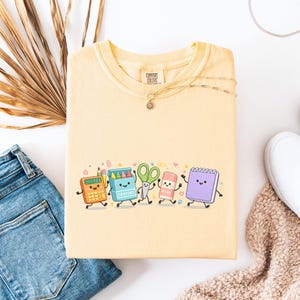 May include: A pale yellow t-shirt featuring a graphic print of anthropomorphic school supplies including a calculator, a box of crayons, green scissors, an eraser, and a purple spiral notebook, all with smiling faces and thin black limbs.