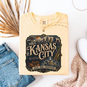 K&ouml;nnte beinhalten: Ein hellgelbes Comfort Colors T-Shirt mit einem Grafikdesign im Vintage-Stil von Kansas City. Das Design zeigt die Skyline der Stadt, ein Saxophon, einen Grill und die Worte "Kansas City, Jazz, BBQ, Fountains."