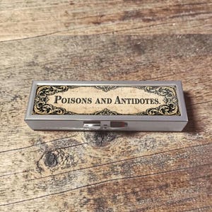 Op de afbeelding: Een rechthoekige, zilverkleurige pillendoos met de woorden "Poisons and Antidotes" in een vintage lettertype. De doos heeft een decoratief, sierlijk randontwerp in zwart en goud. De achtergrond is een houten oppervlak.