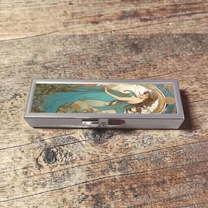Peut inclure: Une boîte à pilules rectangulaire argentée avec un couvercle à charnière. Le couvercle présente une illustration de style Art nouveau d'une femme avec un tissu bleu fluide. Le fond comprend des verts et des bruns sourds.