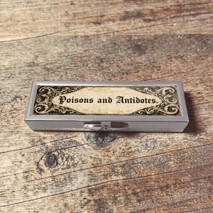 Peut inclure: Une boîte à pilules rectangulaire argentée avec un couvercle à charnière. Le dessus présente une étiquette avec les mots "Poisons and Antidotes" dans une police vintage ornée. L'étiquette a un fond crème avec des volutes décoratives.