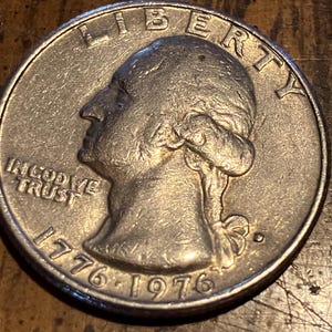 Może przedstawiać: Srebrna moneta o nominale ćwierć dolara z profilem osoby zwróconej w lewo. Moneta zawiera napisy "LIBERTY" i "IN GOD WE TRUST". Widoczna jest również data "1776-1976".