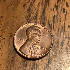 Puede incluir: Una moneda de cobre Lincoln de 1958. La moneda presenta un perfil de Abraham Lincoln, la palabra "LIBERTY" y la frase "IN GOD WE TRUST". También es visible el año "1958".