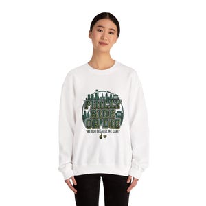Könnte beinhalten: Weißes Sweatshirt mit einem grünen und schwarzen Aufdruck mit der Aufschrift "PHILLY RIDE OR DIE" und einem Stadtbilddesign. Darunter steht der Satz "WE BOO BECAUSE WE CARE" und zwei Herzen.