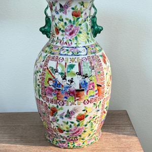 May include: A large porcelain vase featuring a white base with colorful hand-painted floral patterns, birds, and a central panel depicting figures in traditional attire. The vase has two green sculpted handles and a flared rim with yellow trim.