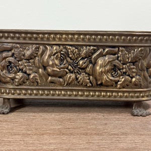 Puede incluir: Una maceta ovalada de latón con un diseño en relieve de rosas florecientes y hojas. El recipiente tiene un borde con cuentas en los bordes superior e inferior y se apoya sobre cuatro pequeños pies detallados en forma de garra en la base.