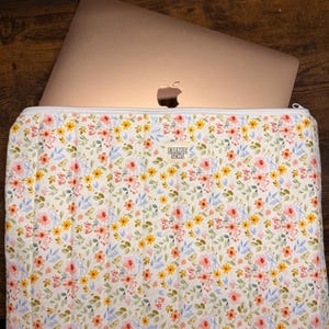 May include: A floral laptop sleeve with a gold laptop inside. The sleeve is white with a pattern of pink, yellow, and blue flowers. The zipper is white, and the brand name "EMMIE SEWS" is printed on the sleeve.