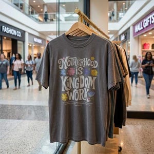 Può includere: Una t-shirt a maniche corte grigio antracite con la scritta bianca Motherhood is Kingdom Work. Il design include illustrazioni floreali colorate che circondano le parole, stampate su un capo a girocollo appeso a una gruccia di legno.