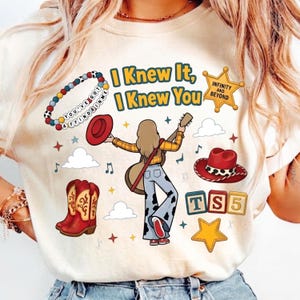 May include: A cream-colored t-shirt featuring a graphic print of a person with a guitar, a red cowboy hat, red boots, a beaded bracelet with the text YOU'VE GOT A FRIEND IN ME, a sheriff star with INFINITY AND BEYOND, and blocks reading TS 5.