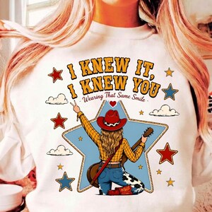 May include: A white sweatshirt featuring a graphic of a person in a red cowboy hat, yellow plaid shirt, and blue jeans holding a guitar. The text reads I KNEW IT, I KNEW YOU, Wearing That Same Smile, surrounded by blue stars, red stars, and clouds.