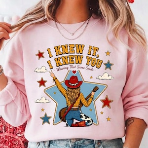 May include: A light pink crewneck sweatshirt featuring a graphic of a person wearing a red cowboy hat, yellow plaid shirt, and cow-print trousers, holding a guitar. The text reads I KNEW IT, I KNEW YOU Wearing That Same Smile, surrounded by stars.